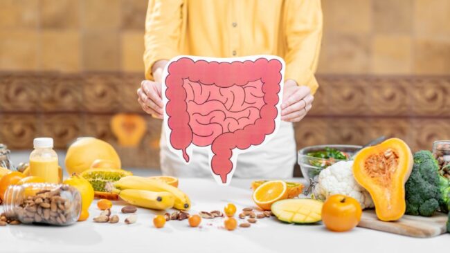 A person holding a pink intestine cutout over a table filled with oranges, broccoli, squash, and nuts.